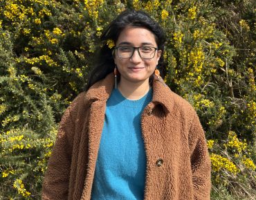 Srijani Datta, a young Asian lady with long black hair, standing outside by a hedge